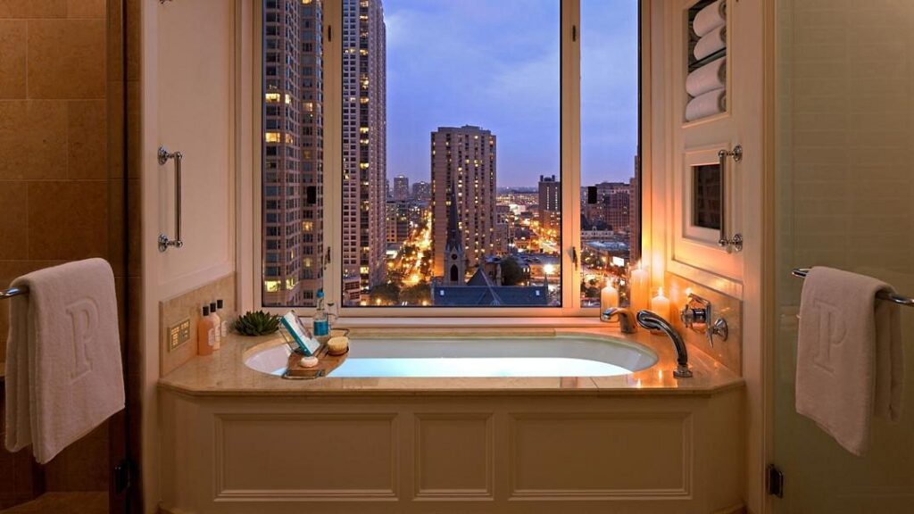 Luxury hotel bathroom with deep soaking tub, city skyline view, candles, and elegant towels at night